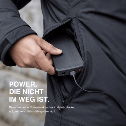 Close-up van een hand die een zwarte powerbank met aangesloten kabel in een jaszak schuift