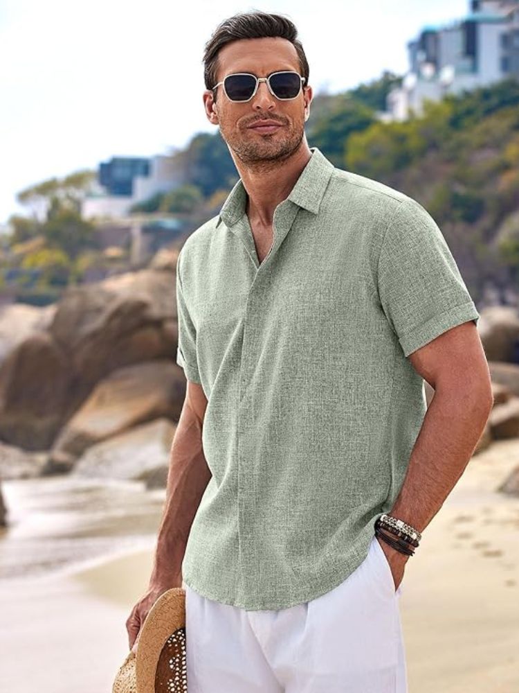Model in mintgroen herenoverhemd en witte broek op het strand, strohoed in de hand