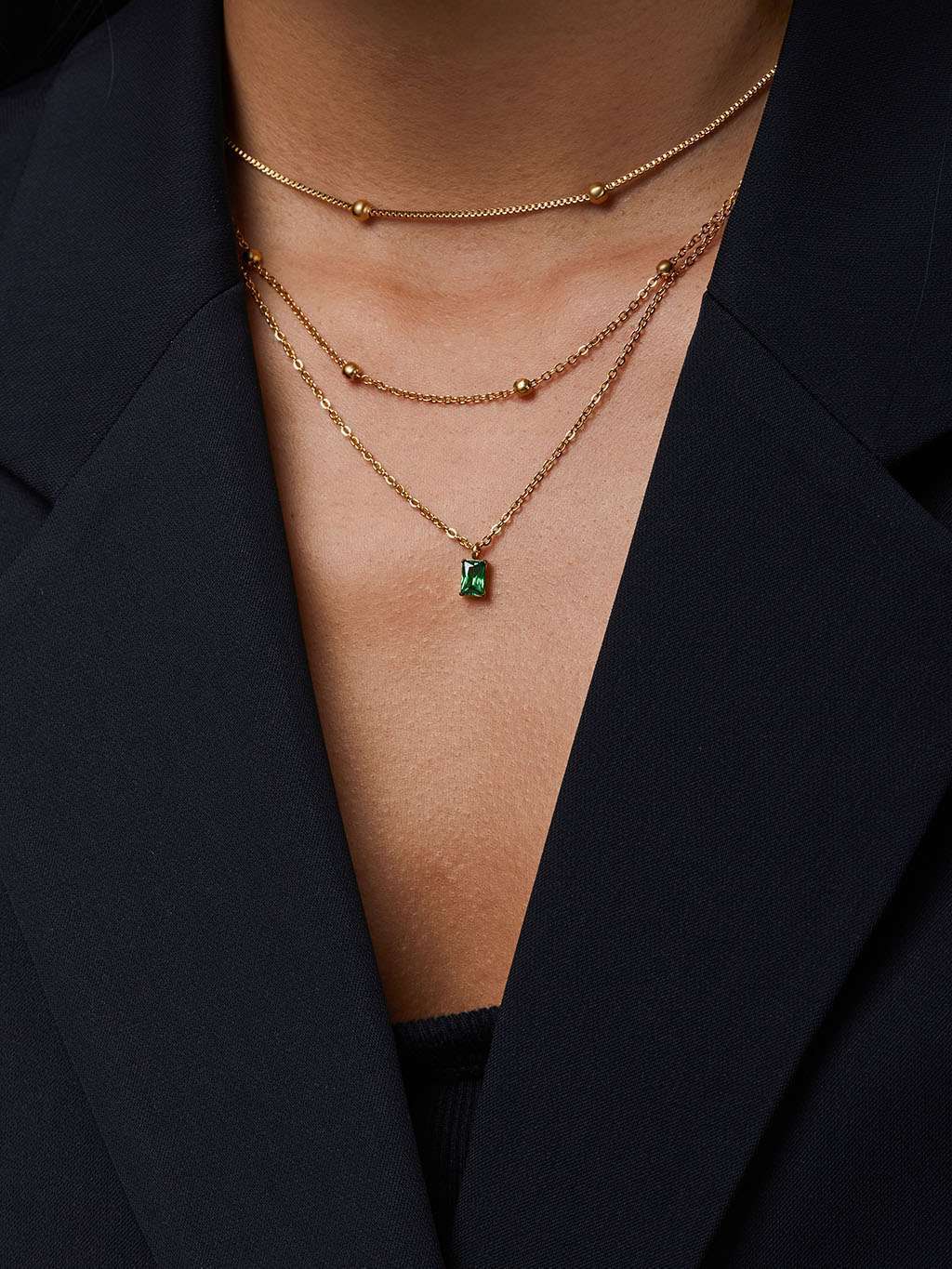 Detail van de ketting met groene hanger gedragen in laagjes met twee andere gouden kettingen onder een zwarte blazer