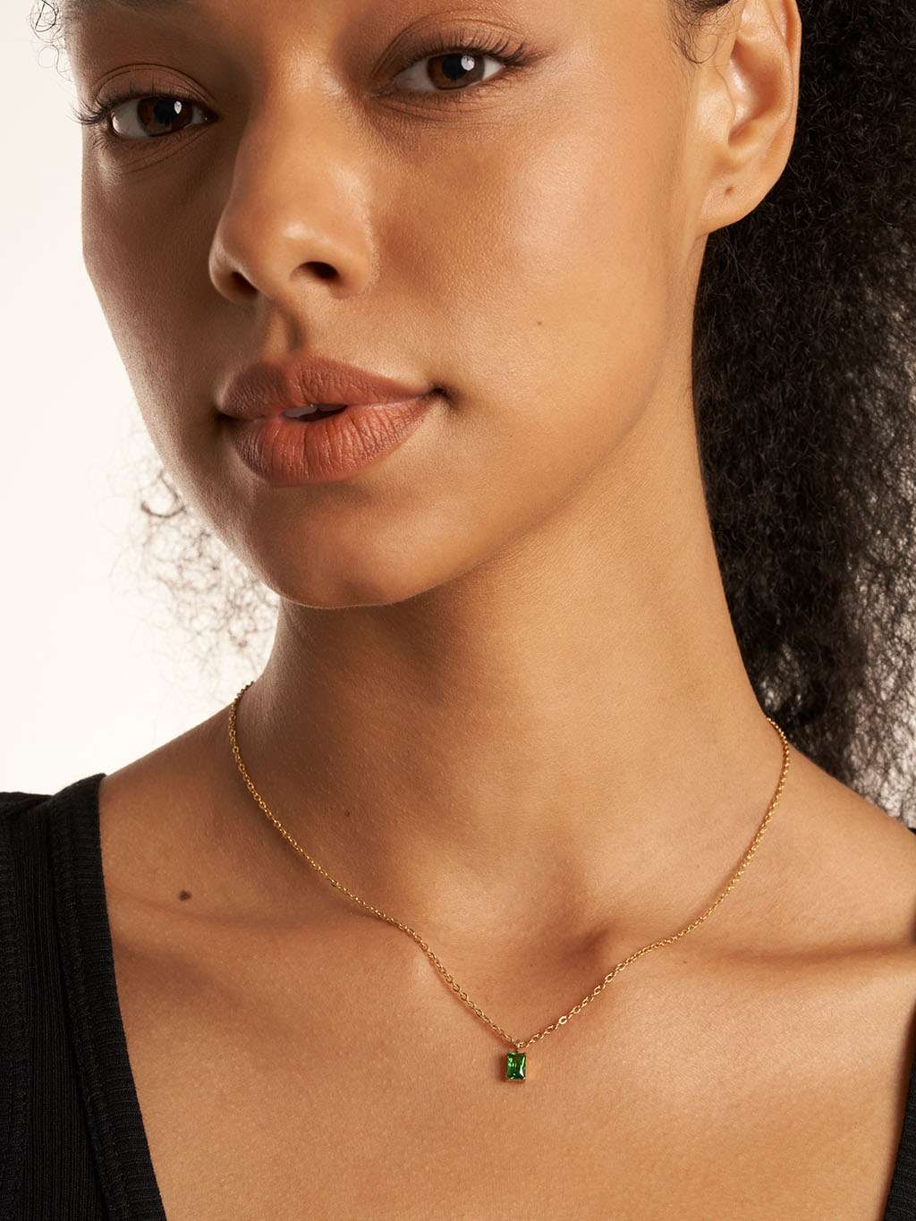 Model met donker krullend haar draagt de gouden ketting met kleine groene hanger op een zwart topje